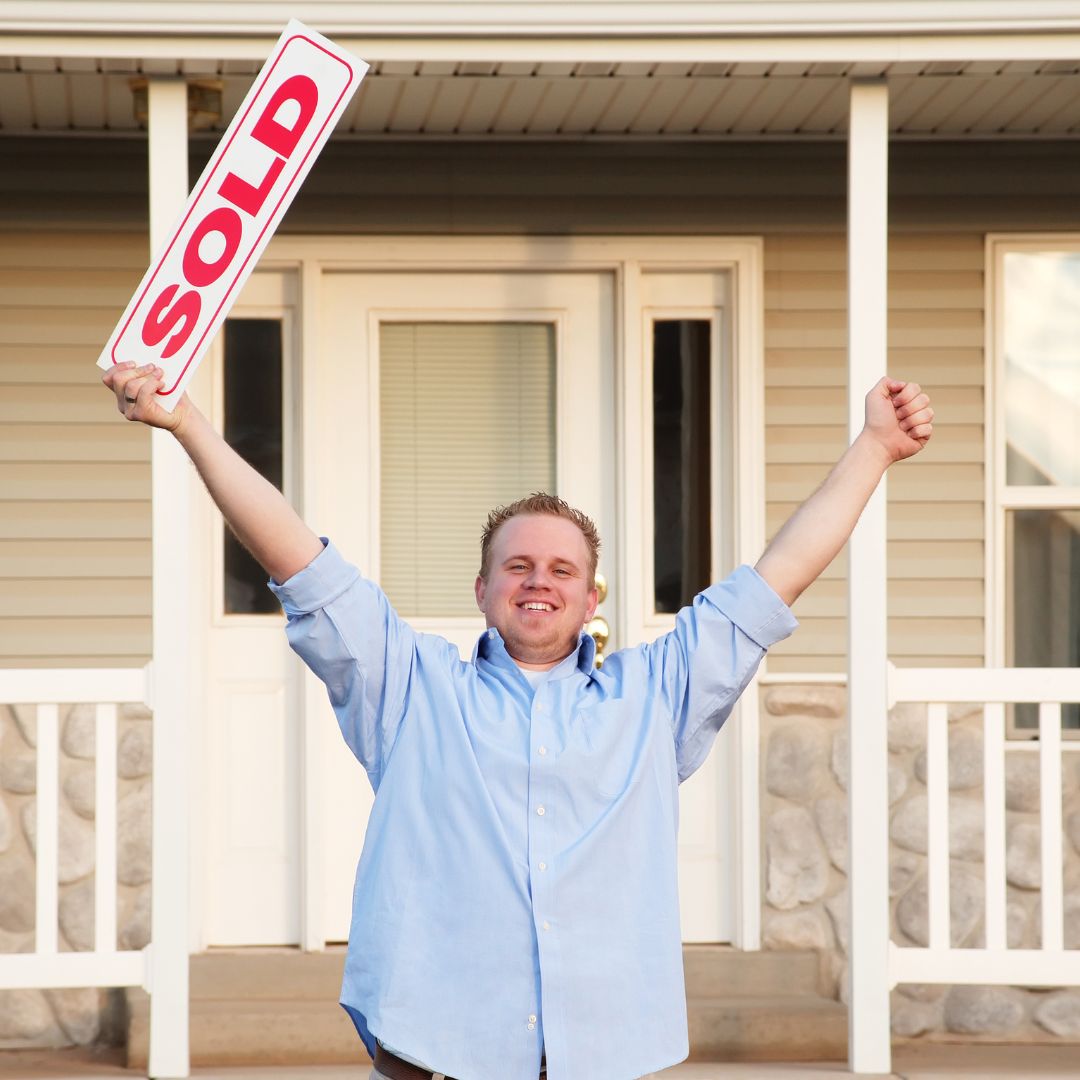 Person in light blue shirt standing on porch raising arms in celebration, holding up a red and white 'SOLD' sign